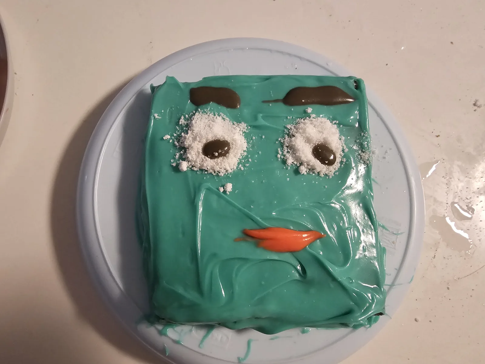 Homemade turquoise square Numberblock Four cake with coconut eyes, chocolate eyebrows, and an orange mouth.