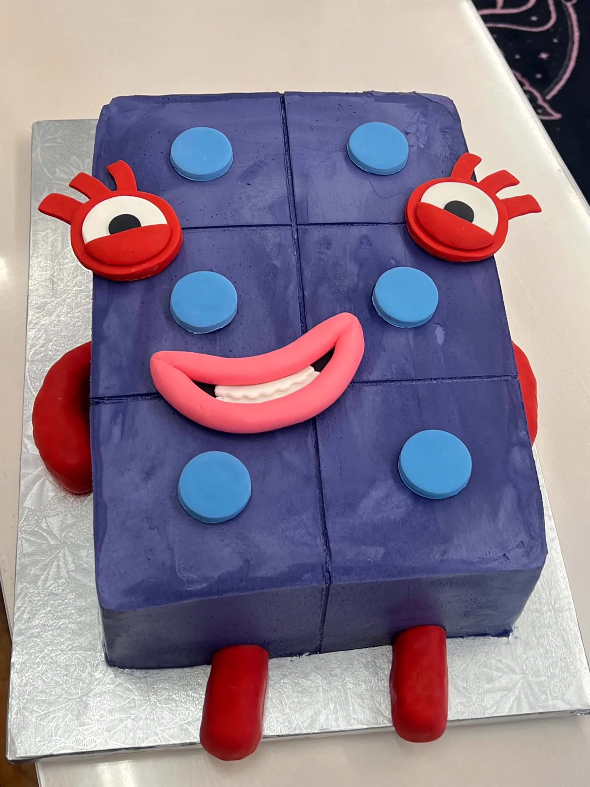 Purple custom Numberblock Six cake made from six square blocks, with red eyes, blue buttons, a pink smile, arms, and legs.