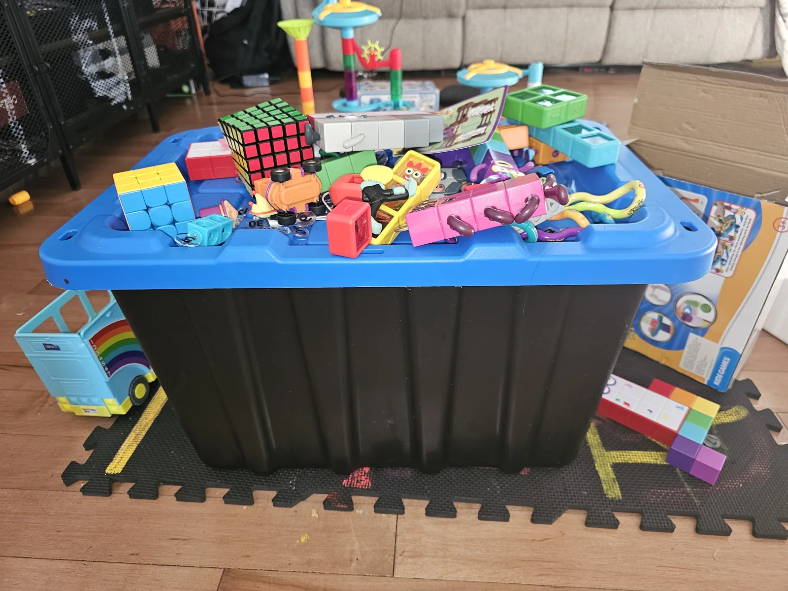 Large black storage box with a blue lid covered in Numberblocks toys, cubes, figures, and games in a living room.